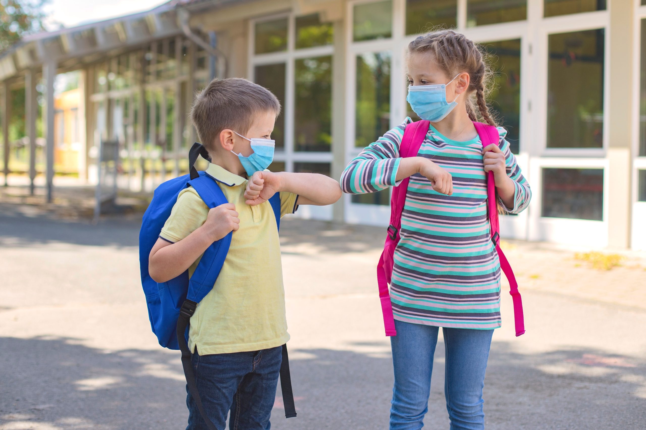 Children, a boy and a girl, wearing masks, greet their elbows on the street in the schoolyard due to the coronavirus epidemic. Distance observance, new school reality.
